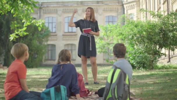 Lección al aire libre para estudiantes de secundaria. Amplio tiro de mujer caucásica positiva de confianza hablando con los alumnos y los niños levantando las manos. Educación al aire libre en un día soleado. — Vídeos de Stock