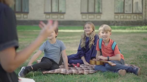 Estudiantes positivos de secundaria que tienen clases al aire libre en un día soleado. Sonrientes chicos y chicas caucásicos levantando las manos y haciendo preguntas como maestro irreconocible haciendo gestos al frente. — Vídeos de Stock