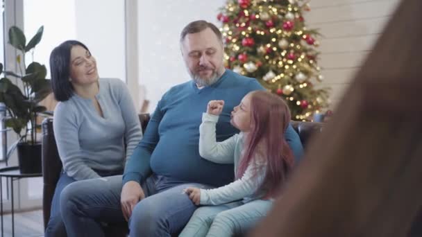 Família feliz sentada no sofá na véspera de Natal e conversando. Retrato de pai, mãe e filha caucasianos felizes que passam férias de Ano Novo em casa. Conceito de celebração. — Vídeo de Stock