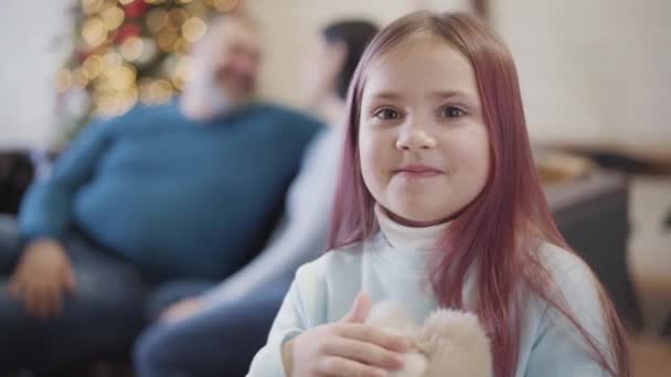 Счастливая симпатичная кавказская девочка, стоящая с игрушкой дома, как родитель, сидящий на диване и разговаривающий на заднем плане. Портрет счастливой дочери, проводящей рождественские каникулы с отцом и матерью дома. — стоковое видео
