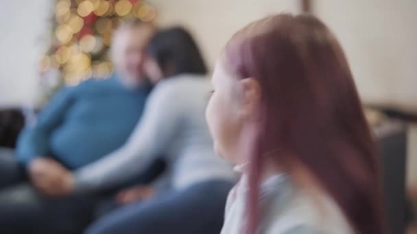 Câmera segue linda menina correndo para os pais sentados no sofá com a árvore de Ano Novo no fundo. Feliz filha caucasiana desfrutando de fins de semana de Natal com pai e mãe em casa. — Vídeo de Stock