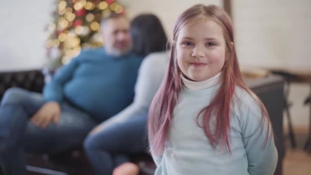 Vidám kaukázusi lány a kamerába néz, és a boldog szülőkhöz rohan, akik a kanapén ülnek a háttérben. Bájos lány élvezi a karácsonyi ünnepeket otthon. Rögzítés. — Stock videók