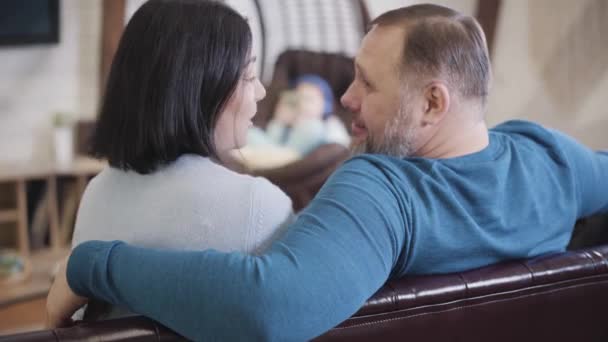 Visão traseira de amor médio adulto casal caucasiano beijando como criança borrada sentado em cadeira pendurada no fundo. A disparar por cima do ombro. Família feliz desfrutando de fins de semana juntos em casa. — Vídeo de Stock