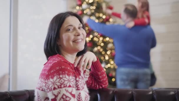 Porträt einer glücklichen Frau, die in die Kamera lächelt und auf verschwommene Männer und Töchter zurückblickt, die den Weihnachtsbaum schmücken. Idyllische Neujahrsferien der kaukasischen Familie. — Stockvideo
