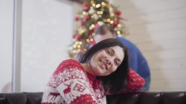 사랑하는 딸과 남편이 소파에 앉아 크리스마스 트리 장식을 하고 웃고 있는 아름다운 여성을 껴안고 있습니다. 행복 한 새해 휴일 백인 가족의 긍정적 인 휴일. — 비디오