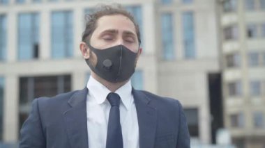 Covid-19 yüz maskeli stresli adam ofis binasının önünde el ele tutuşarak dikiliyor. Coronavirus salgını sırasında sorun yaşayan Kafkas işadamının portresi.