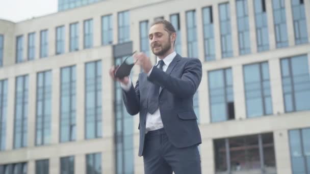 Happy Caucasian businessman in formal suit taking off Covid-19 face mask and throwing it away. Portrait of excited young man rejoicing end of coronavirus pandemic, business building at the background. — Stock Video