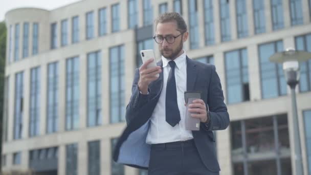 Retrato de un hombre de negocios nerd concentrado utilizando el teléfono inteligente al aire libre en frente del edificio de negocios de oficinas. Enfocado rico hombre caucásico en anteojos navegar por Internet en coffee break. — Vídeos de Stock