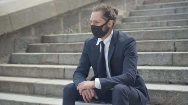 Retrato de un empresario deprimido con exceso de trabajo en Covid-19 máscara de la cara sentado en las escaleras urbanas de la ciudad y suspirando. Joven hombre caucásico en traje formal pensando al aire libre en la pandemia de coronavirus. — Vídeos de Stock