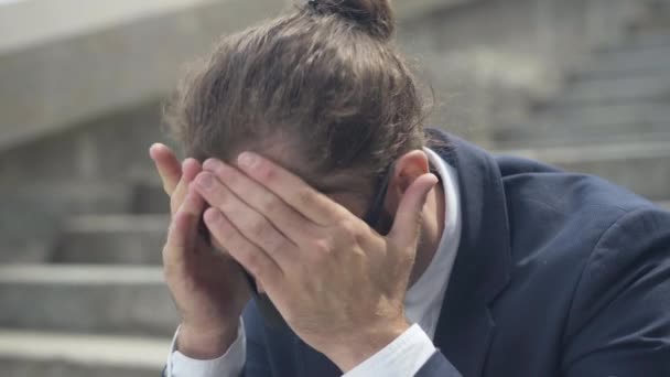 Närbild av deprimerad man i Covid-19 ansiktsmask håller huvudet med händer och tänkande. Porträtt av stressad kaukasisk affärsman sitter på soliga urbana trappor utomhus under coronavirus pandemi. — Stockvideo