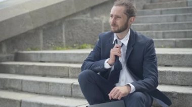 Üzgün iş adamı kravatını gevşetiyor ve gömlek düğmesini açıyor. Şehir merdivenlerinde oturup düşünen mutsuz beyaz bir gencin portresi. Dışarıdaki bitkin girişimci..