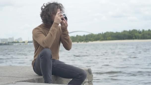 Kreativer gutaussehender Mann sitzt mit Retro-Kamera am Ufer und macht Fotos. Porträt eines selbstbewussten kaukasischen Fotografen, der einen düsteren Abend im Freien mit verschwommener Stadt im Hintergrund verbringt. — Stockvideo