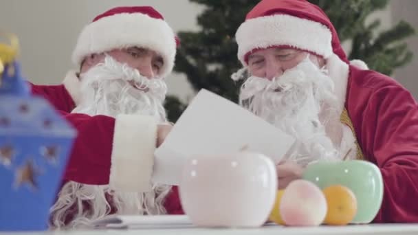 Dois Pai Natal sentados à mesa a ler cartas. Retrato de homens caucasianos jovens e seniores em trajes e barbas falsas no Ano Novo ou Natal. Feriados e celebração conceito. — Vídeo de Stock