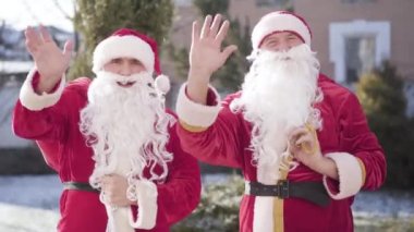 Genç ve yaşlı Noel Babalar dışarıda kameraya el sallıyorlar. Mutlu Noeller dileyen iki neşeli Noel Baba 'nın portresi. Yeni yıl kutlaması ve tatil sezonu.