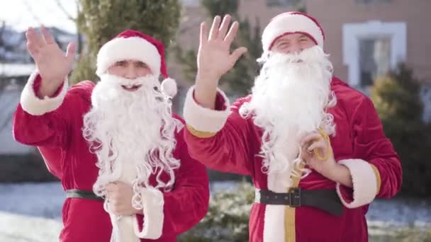 Santas jóvenes y mayores de pie al aire libre y saludando a la cámara. Retrato de dos alegres Santa Claus deseando feliz Navidad. Celebración de Año Nuevo y temporada de fiestas. — Vídeo de stock