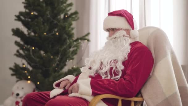 Cansado Santa Cláusula sentado em cadeira de balanço com árvore de Natal decorada no fundo. Jovem exausto em traje vermelho adormecendo na véspera de Ano Novo. Temporada de férias. — Vídeo de Stock