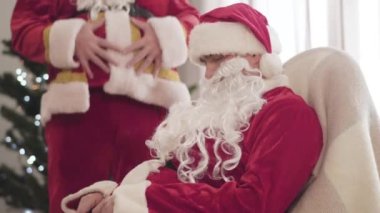 Kırmızı kostümlü, sallanan sandalyede uyuyan genç adam arka planda karnını tutarak gülen tanınmamış bir Noel Baba. Noel arifesinde eğlence. Yeni yıl kutlaması.