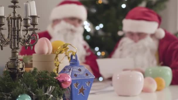 Decoraciones de Navidad de pie sobre la mesa como dos Santa Claus borrosas hablando en el fondo. Santas irreconocibles leyendo cartas en Nochevieja. — Vídeo de stock