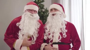 Kırmızı kostümlü, gülen ve göbek tutan iki Noel Baba portresi. Noel arifesinde arka planda yılbaşı ağacıyla evde eğlenen eğlenceli beyaz adamlar..