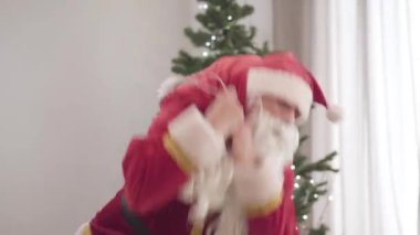 Yaşlı Noel Baba 'nın kırmızı kostümlü portresi ağır hediye torbalarını kaldırıyor. Yaşlı beyaz adam elinde Noel hediyeleriyle kırmızı çanta tutuyor. Yeni yıl ağacı arka planda duruyor..