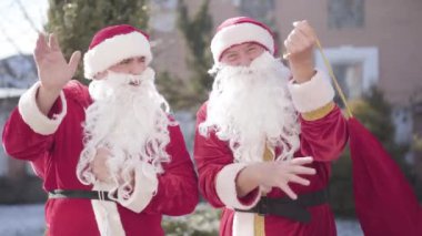 Kırmızı Noel Baba kostümlü iki pozitif adam kırmızı hediye torbalarını açıp sallıyor. Neşeli gülümseyen Noel Babaların portresi Noel arifesinde Noel hediyeleri getiriyor..