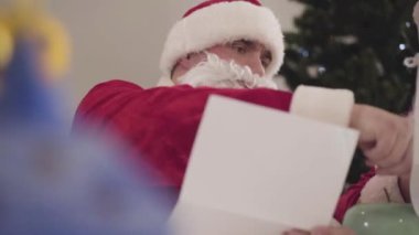 Kırmızı Noel Baba kostümlü genç beyaz adamın portresi mektupları okuyor. Noel arifesinde ciddi bir Noel Baba. Arka planda yeni yıl ağacı..