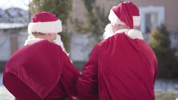 Δύο Αϊ-Βασίληδες με Χριστουγεννιάτικα κοστούμια με σακιά δώρων να γυρίζουν, να χαιρετάνε την κάμερα και να φεύγουν. Πίσω όψη του Άγιου Βασίλη περπατώντας μακριά με κόκκινες σακούλες. Παραμονή Πρωτοχρονιάς. — Αρχείο Βίντεο