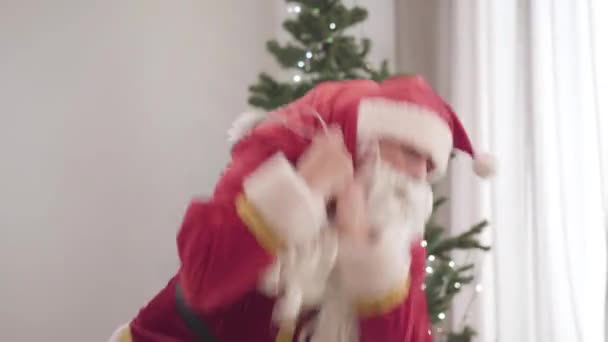 Retrato de Santa idosos em traje vermelho levantando pesados presentes saco. Homem caucasiano velho segurando saco vermelho com presentes de Natal. Árvore de Ano Novo decorada em pé no fundo dentro de casa. — Vídeo de Stock