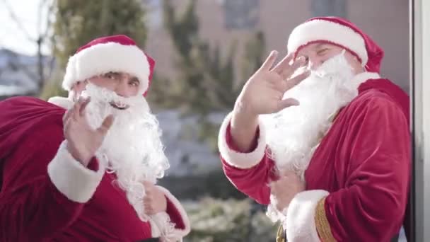 Dva pozitivní Santové mávají před kamerou, plácají si a odcházejí. Portrét usmívajícího se veselého bělocha v červených novoročních kostýmech na Štědrý den. — Stock video