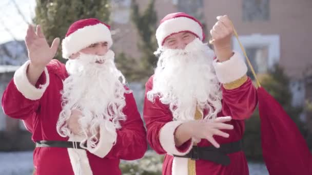 Két pozitív férfi piros Mikulás jelmezben integet és kinyitja a piros ajándékzsákot. Portré vidám mosolygós Mikulások hozza karácsonyi ajándékokat szilveszterkor. — Stock videók