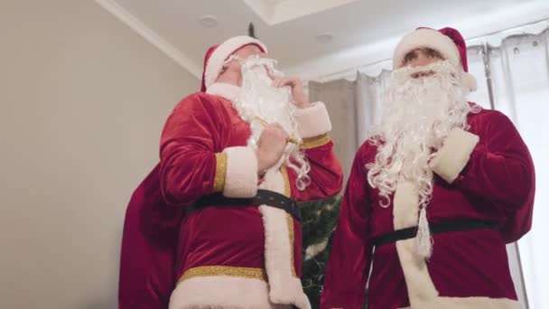 身穿红色圣诞老人服装的年轻和年长的白人男子的画像，摘下假胡子，叹了口气。累了的圣诞老人带着礼品袋在屋里聊天圣诞节庆祝活动. — 图库视频影像