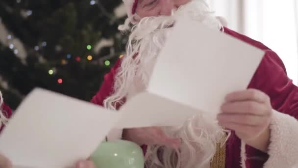 シニアサンタは赤いクリスマスの衣装の若者と手紙を読んでいます。深刻なサンタ句の肖像新年の前夜に若い同僚を教える. — ストック動画