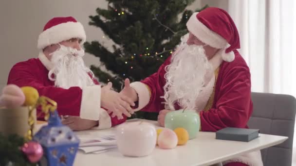 圣诞前夕,年轻的和年长的圣诞老人握手.两个穿着圣诞老人服装的自信男子坐在桌旁，与圣诞树握手的肖像. — 图库视频影像