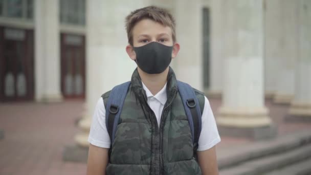 Uczeń w masce Covid-19 patrzy w kamerę i idzie do budynku szkoły. Portret poważnego chłopca studiującego podczas pandemii koronawirusowej. Styl edukacji Covid. — Wideo stockowe