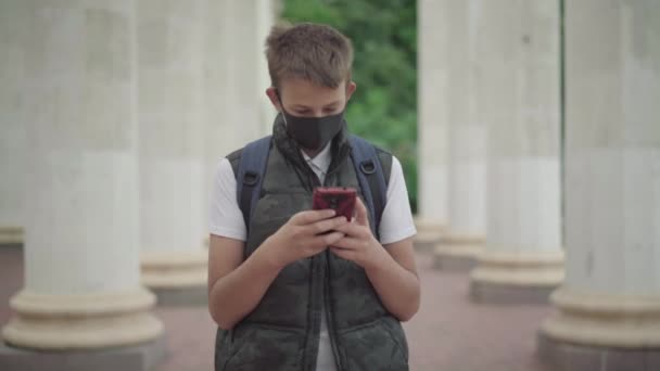 Covid-19 maskeli beyaz bir öğrenci dersten sonra dışarıda yürüyor ve akıllı telefondan internette sörf yapıyor. Coronavirus salgını sırasında okuldan sonra internetten mesajlaşan çocuğun portresi. — Stok video