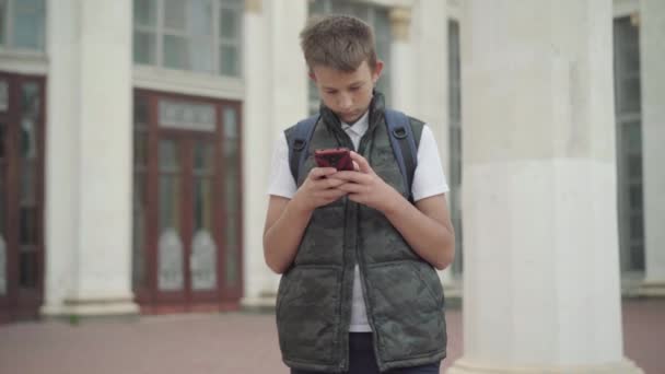 Portret van een boeiende schooljongen die online berichten verstuurt. Geabsorbeerde blanke jongen met behulp van sociale media op smartphone buiten staan. Generatie Z Internetverslaving. — Stockvideo