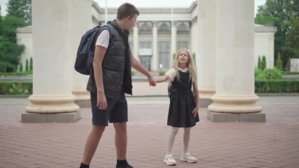 Široký portrét školáka, jak tahá za ruku tvrdohlavou školačku, která se nechce jít učit. Roztomilá sestra stojící a mluvící jako bratr přesvědčující ji, aby šla do školy. — Stock video
