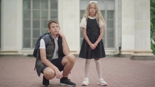 Stubborn schoolgirl standing and talking as elder brother persuading her to go to school. Portrait of Caucasian siblings outdoors before lessons. Public education. — Stock Video