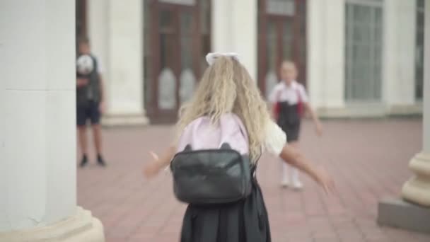 Ragazza carina correre e abbracciare compagno di classe dopo le vacanze scolastiche. Ragazzo sfocato in piedi con pallone da calcio sullo sfondo. Felice scolari incontro prima delle lezioni all'aperto. — Video Stock