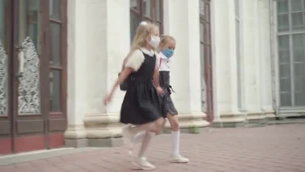 Камера слідує за веселими кавказькими дівчатами, які бігають і стрибають за класами під час пандемії коронавірусу. Дві радісні школярки в Ковід-19 стикаються з масками веселощів після навчання.. — стокове відео