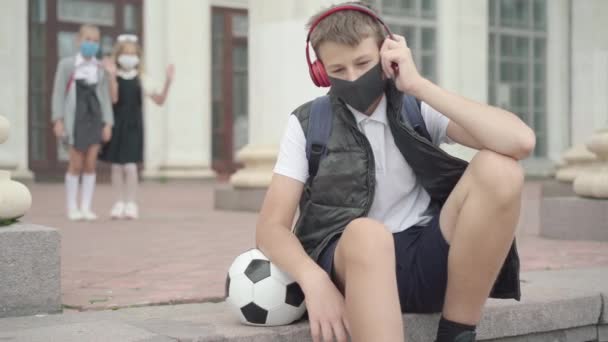 Relaxed Caucasian schoolboy in Covid face mask sitting on school stairs with sport ball and listening to music in headphones. Blurred schoolgirls chatting at the background. Classmates resting. — Stock Video