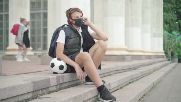 Wide shot of relaxed schoolboy sitting on school stairs and listening to music in headphones as schoolgirls passing at the background. Portrait of carefree boy resting after studying outdoors. — Stock Video