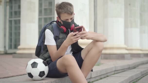 Ragazzo caucasico in maschera facciale seduto sulle scale della scuola con pallone da calcio e internet surf su smartphone. Ritratto di scolaro che riposa dopo le lezioni durante la pandemia di Covid. — Video Stock