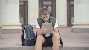 Neşeli kızlar okul merdivenlerinde oturan ve tablet kullanan bir çocuğa koşuyorlar. Sınıf arkadaşları dışarıda çalıştıktan sonra dinleniyor. Neşeli çocuklar dersten sonra eğleniyorlar..