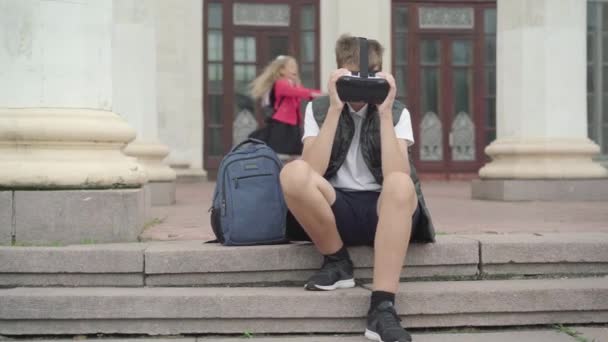 Niño caucásico usando auriculares de realidad virtual como chicas borrosas girando en el fondo. Retrato de escolares despreocupados jugando mientras las colegialas disfrutan de las vacaciones escolares al aire libre. — Vídeos de Stock