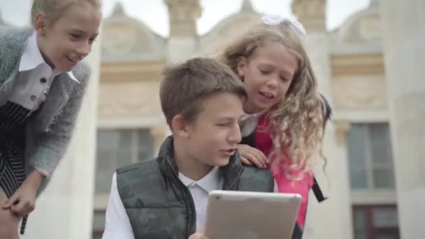 Trois écoliers caucasiens joyeux surfant sur Internet sur tablette pendant la pause scolaire. Portrait de garçons et de filles positifs bavardant et utilisant les médias sociaux à l'extérieur après avoir étudié. — Video