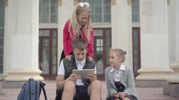 Charmante Kinder sitzen in der Pause draußen mit Tablet und unterhalten sich. Porträt glücklicher Jungen und Mädchen, die nach dem Studium in der Schule ruhen. Bildungs- und Freizeitkonzept. — Stockvideo