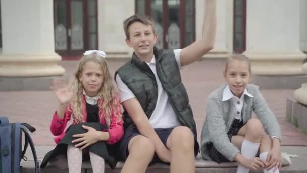 Três crianças positivas acenando e sorrindo para a câmera. Retrato de meninas caucasianas alegres e menino descansando depois de estudar ao ar livre. Crianças em idade escolar se divertindo depois das aulas. — Vídeo de Stock