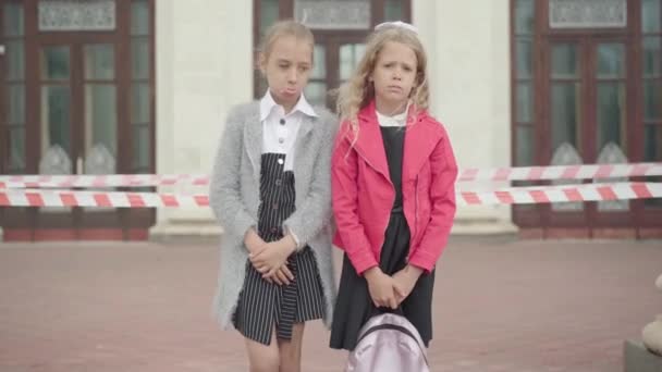 Portret van ontevreden blanke schoolmeisjes die voor school stonden, gesloten met barricade tape en kijkend naar de camera. Trieste meisjes ongelukkig met de educatieve onderbreking tijdens de coronavirus pandemie. — Stockvideo