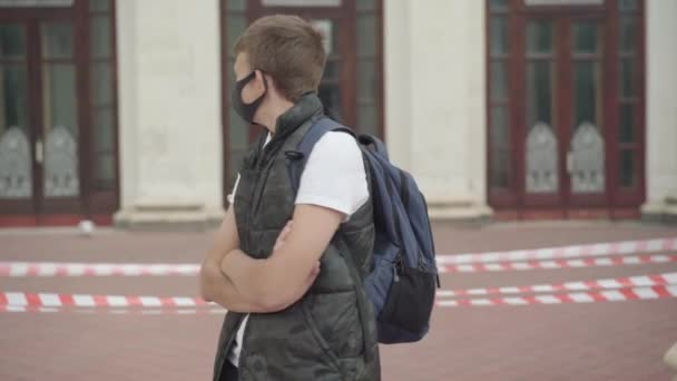 Upprörd pojke i Covid ansiktsmask som står med händerna korsade och ser sig omkring. Porträtt av sorgsen vit skolpojke framför stängd skola utomhus omgiven av barrikadtejp. — Stockvideo